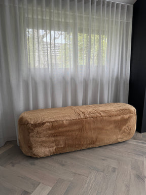 Brown textured bench on a wooden floor with sheer curtains in the background