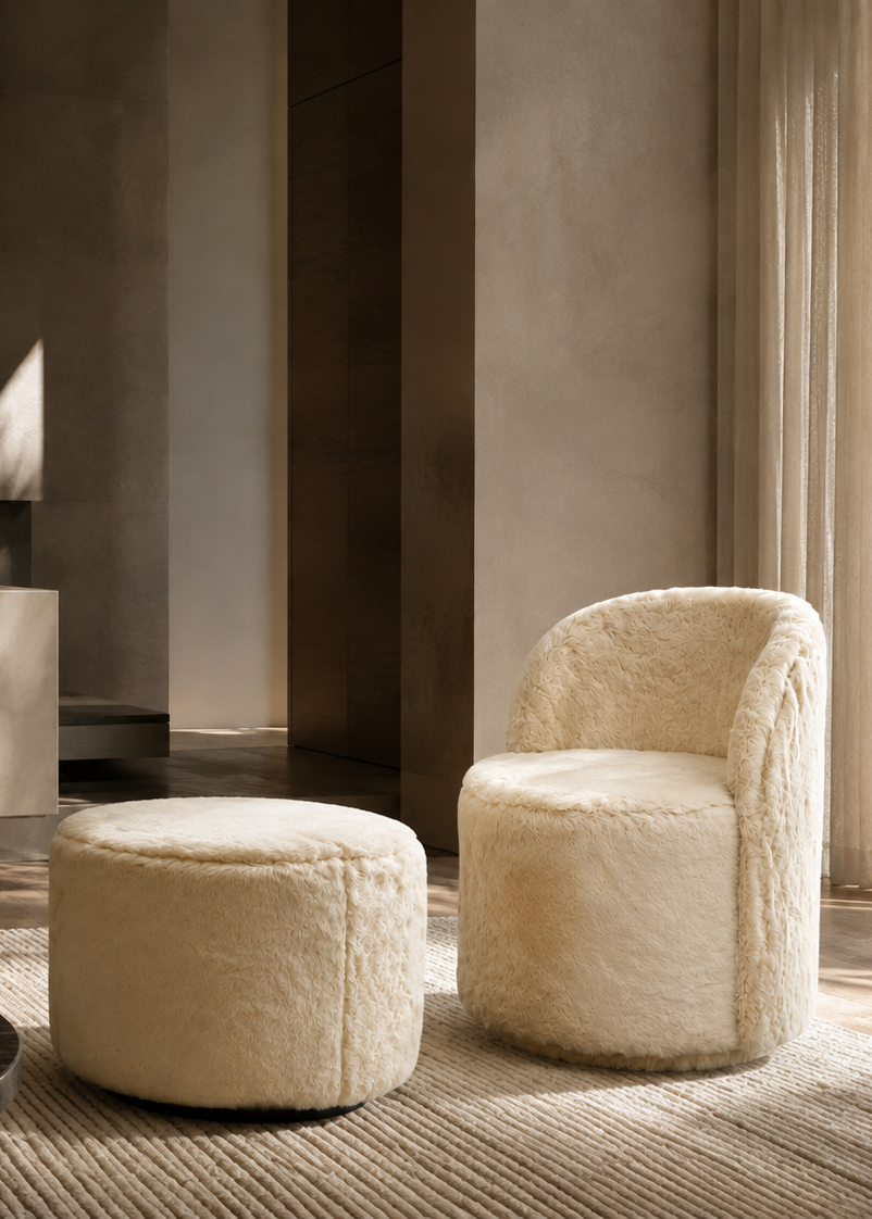 A modern cream-colored sheepskin armchair and matching round ottoman in a minimalist, warm-toned living room setting.