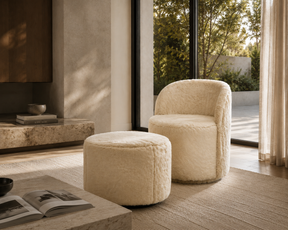 A cream faux fur armchair with a matching round ottoman in a modern, sunlit living room with large windows and a stone fireplace.