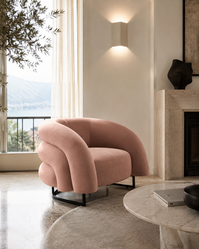 A blush pink velvet accent chair with a curved, sculptural backrest and black metal legs, positioned in a modern living room next to a marble fireplace and a large window with an outdoor view.