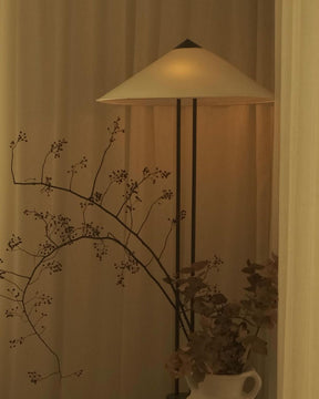 Floor lamp with a beige shade in a dimly lit room, next to a plant and branches.